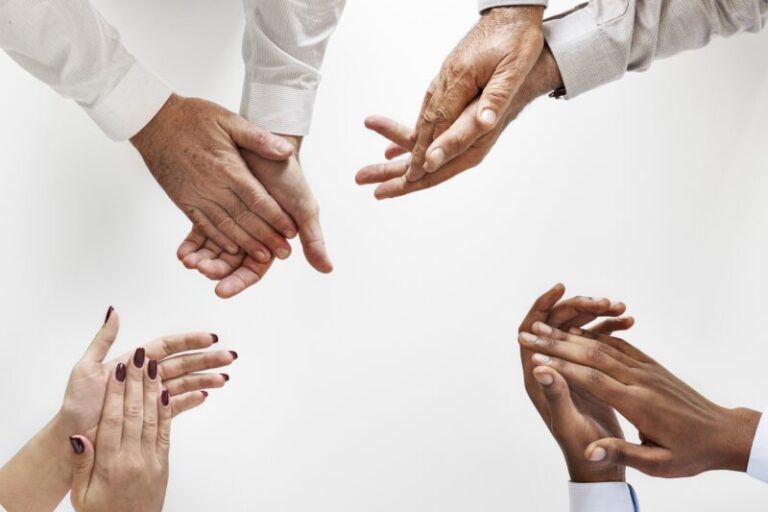 overhead view of four pairs of hands in clapping position, white shirts shown on arms - image is cropped at wrists